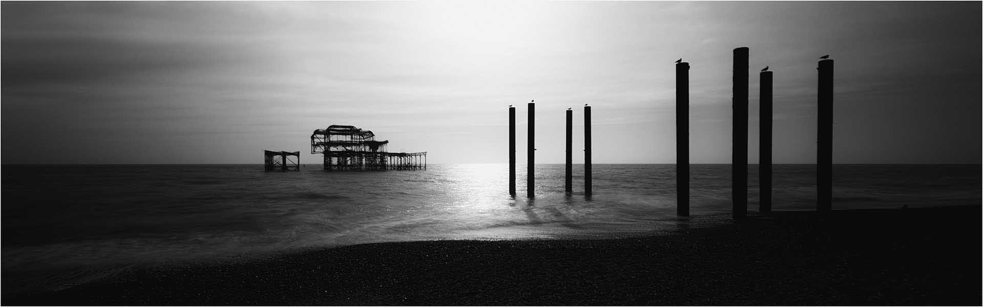 Minimalist black-and-white seascape with calm water, silhouetted posts, and a skeletal pier structure on the horizon.
