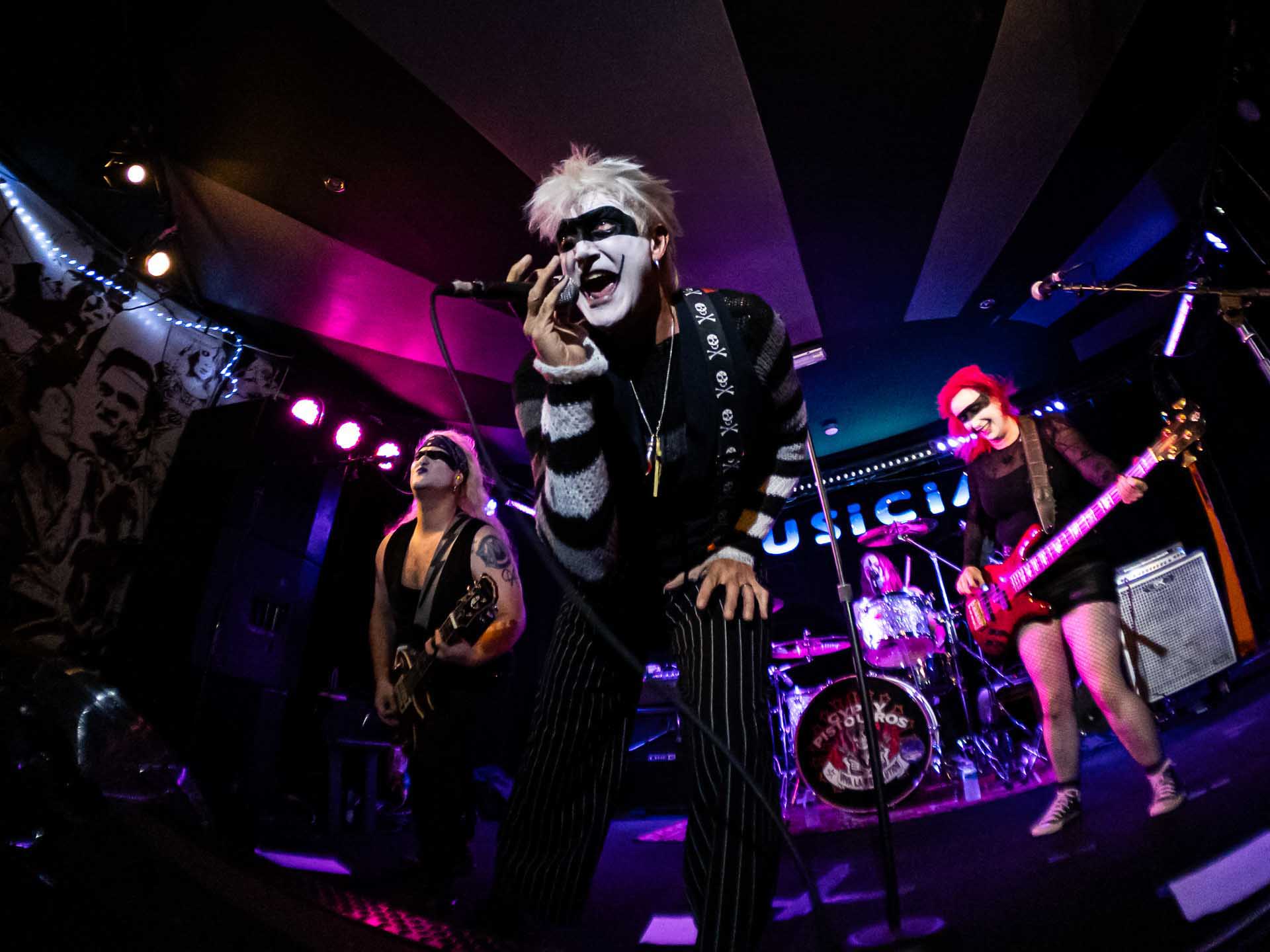 Rock band performing on stage under colorful lights, lead singer in face paint singing into a microphone while guitarists play.