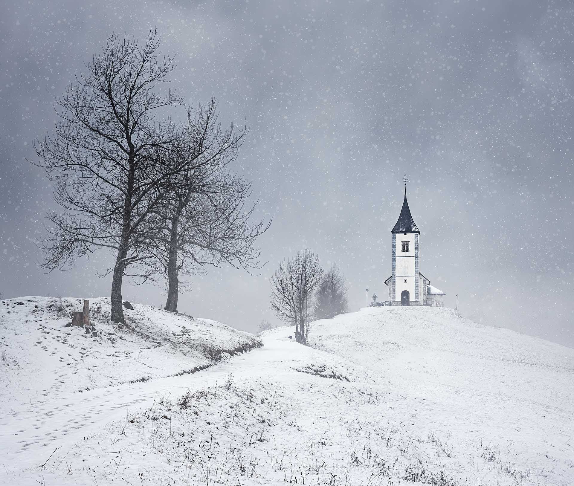 Snowy landscape with a small church on a hill, bare trees, and a winding path during falling snow.