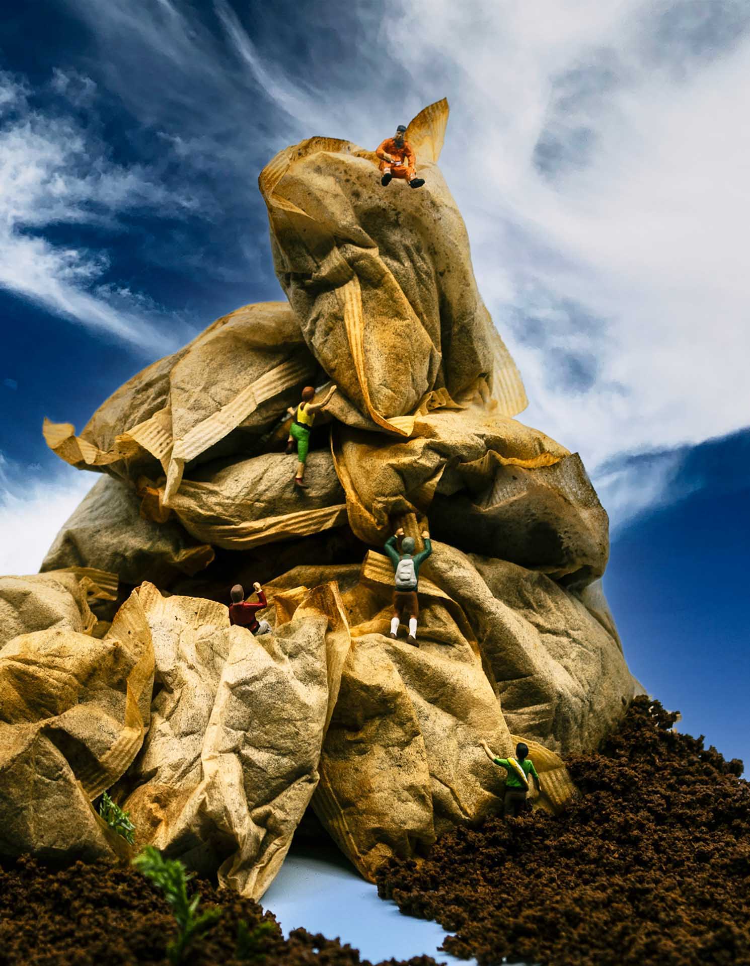 Miniature climbers scaling a crumpled paper mountain under a blue sky, creating a playful illusion.