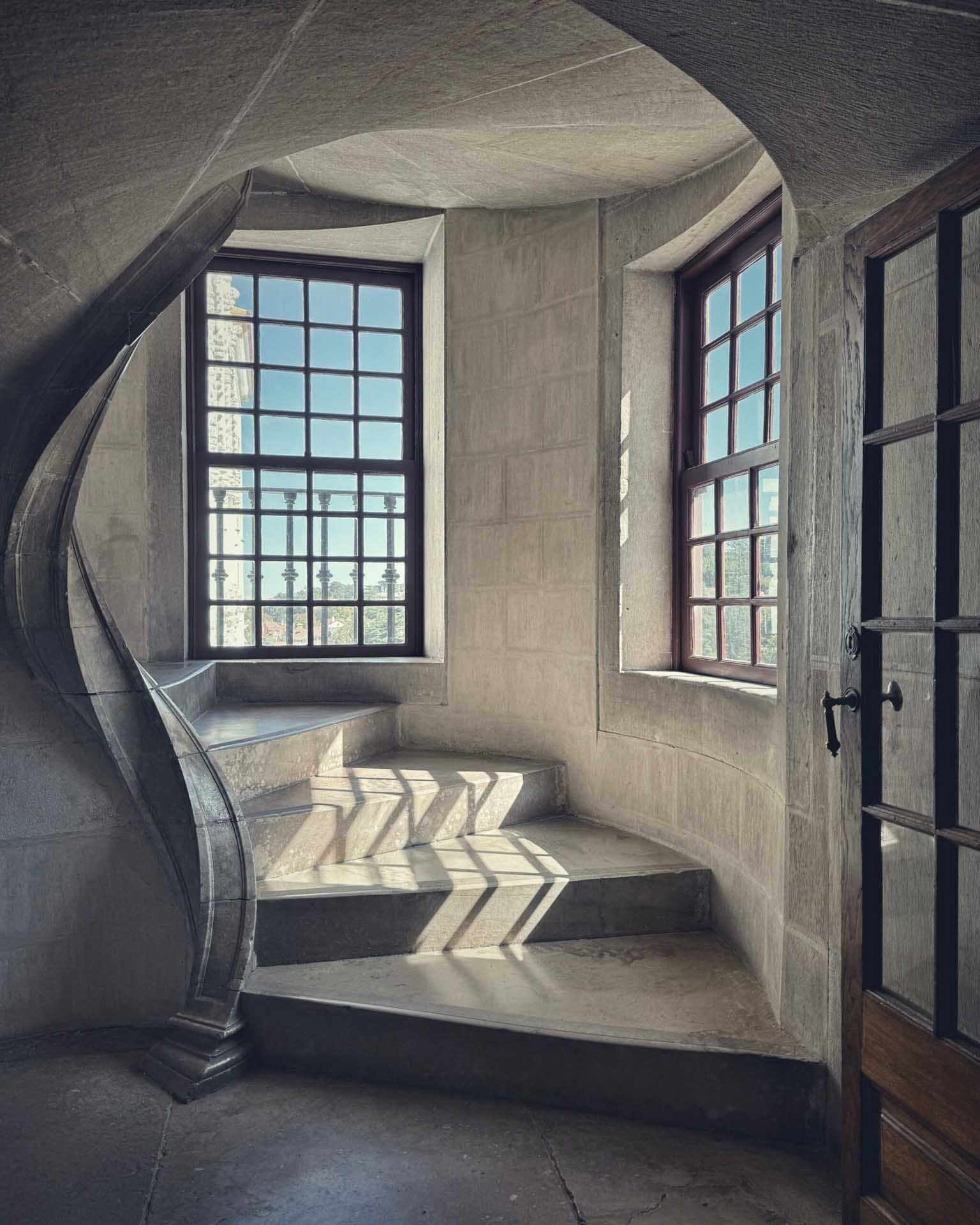 Curved stone staircase inside a building with sunlight casting patterned shadows through large windows.