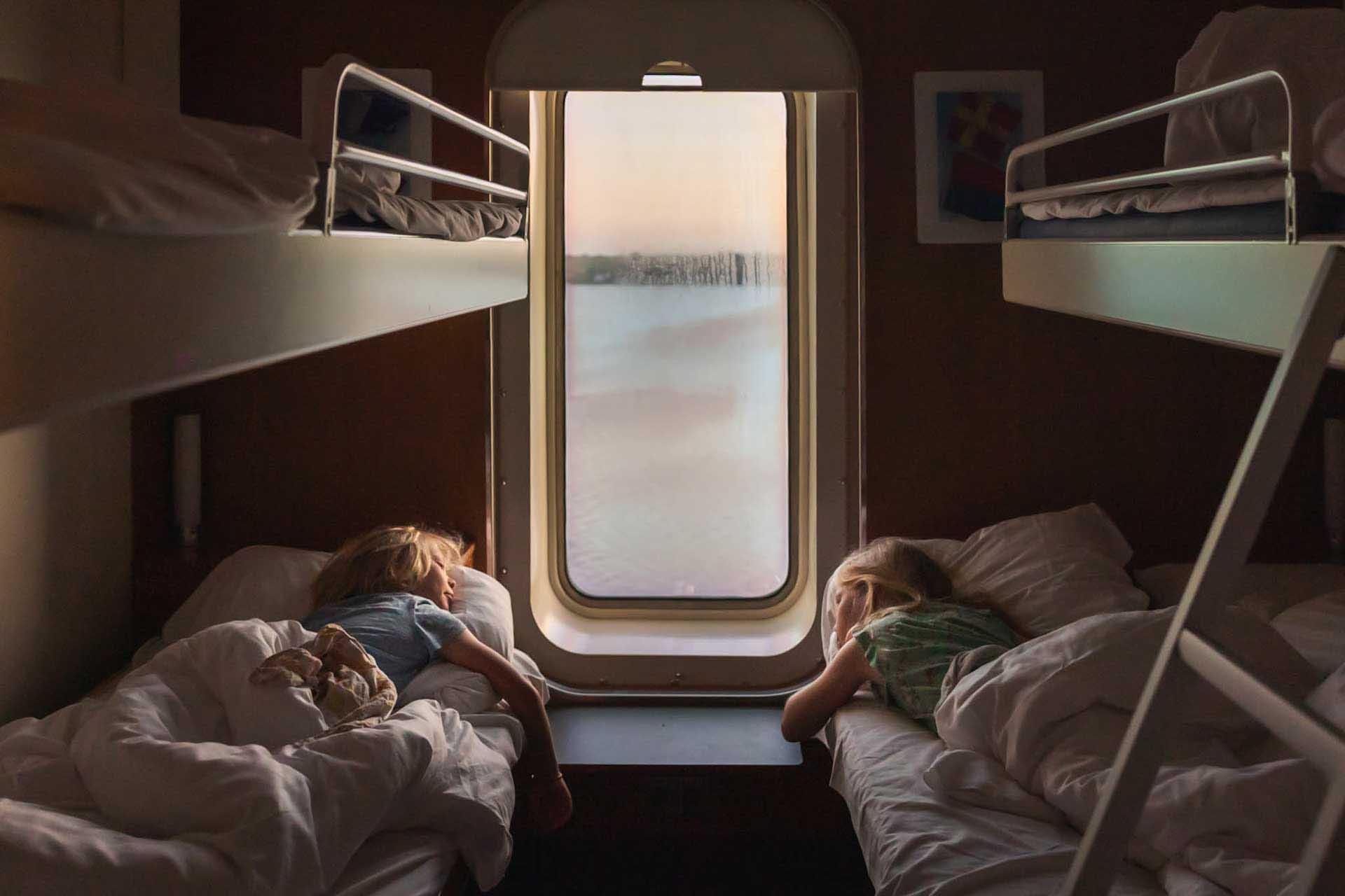 Two children sleeping in bunk beds inside a train cabin with a window showing blurred scenery outside.