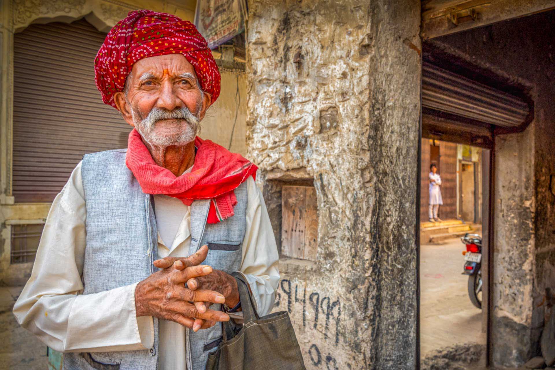 Elderly man wearing a red turban and scarf standing in a weathered street setting with textured walls and a doorway behind him.