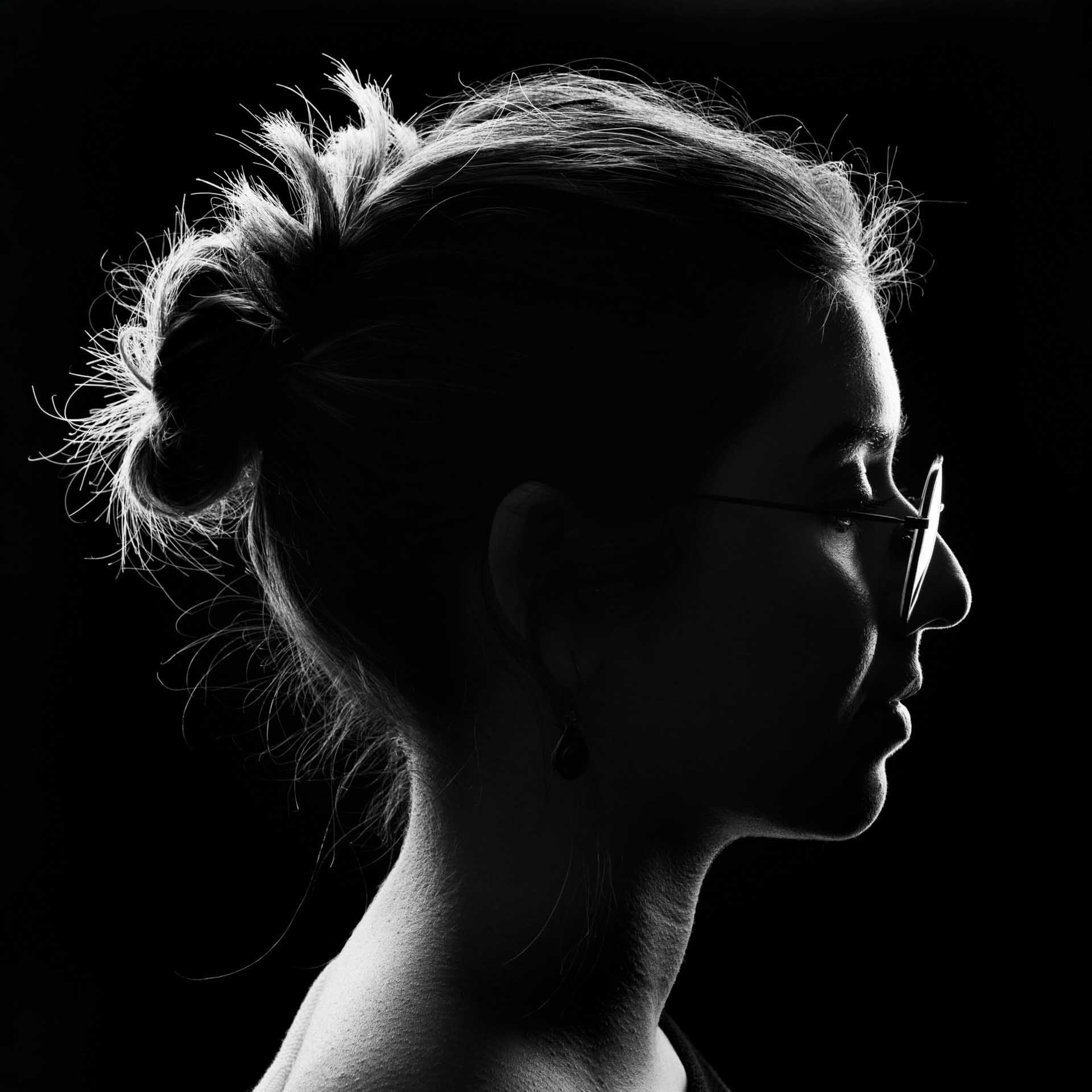 Black-and-white side profile silhouette of a person with hair in a bun and glasses against a dark background.