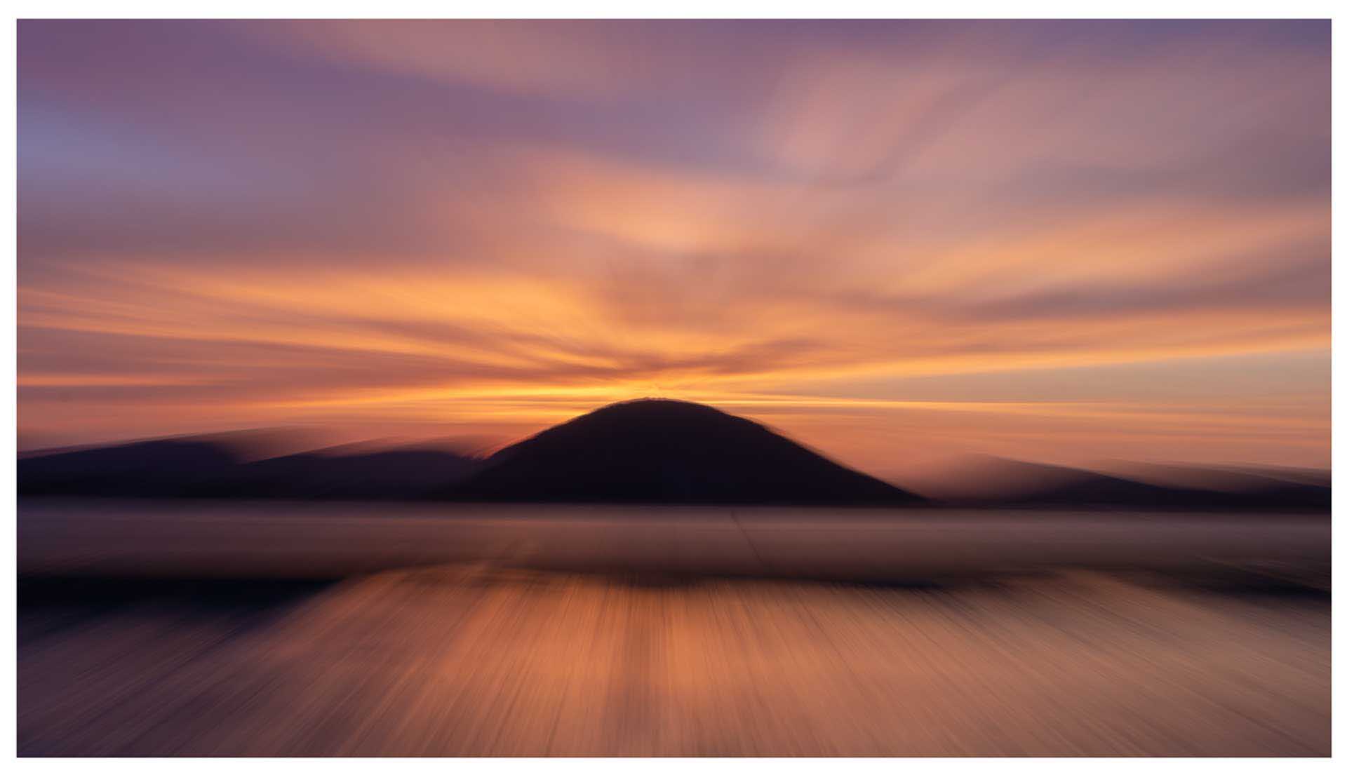 Soft-focus landscape with a hill silhouetted against a colorful sunset sky in warm tones.