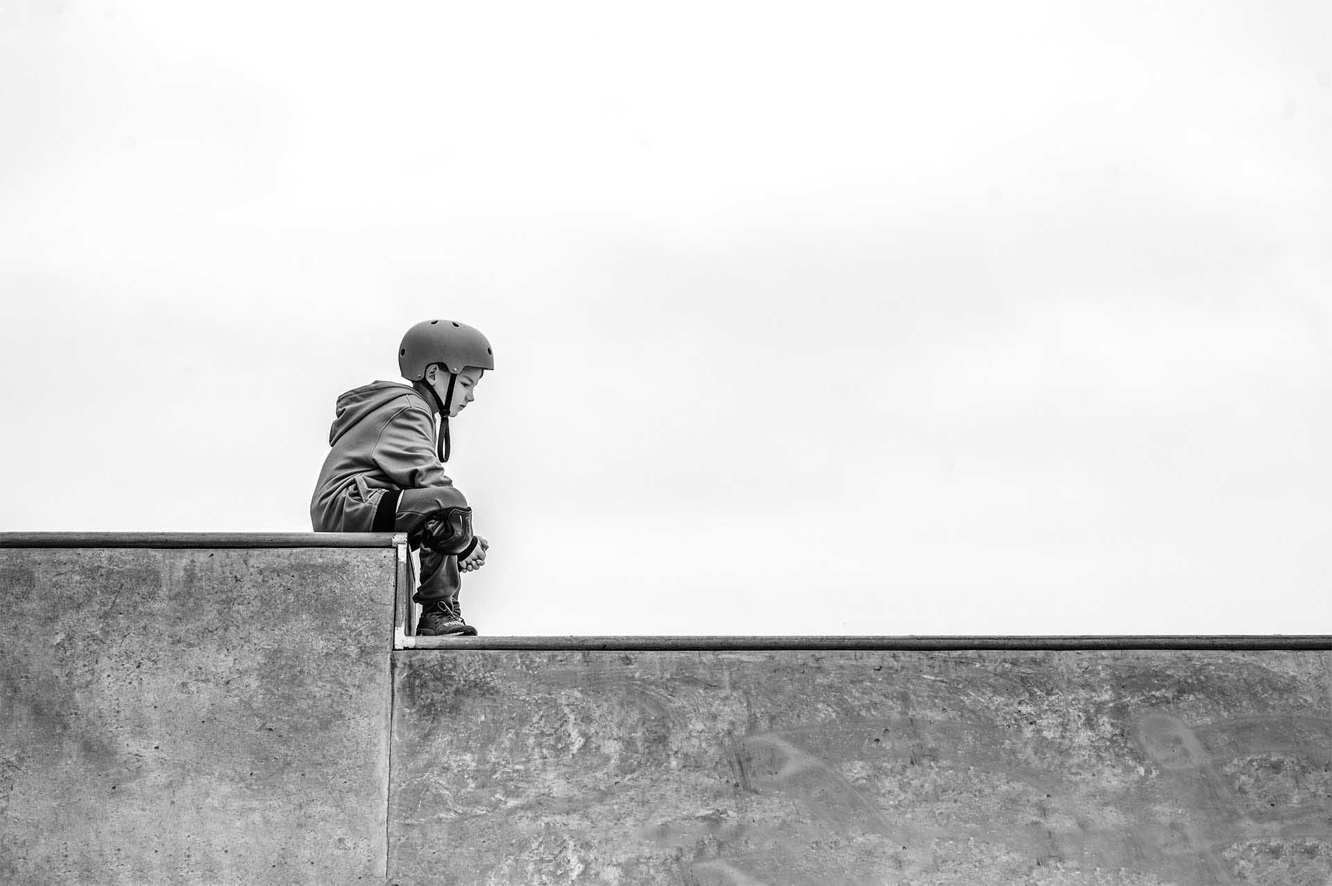 Black-and-white photo of a child wearing a helmet sitting on the edge of a concrete skate ramp, looking down with large empty space around.
