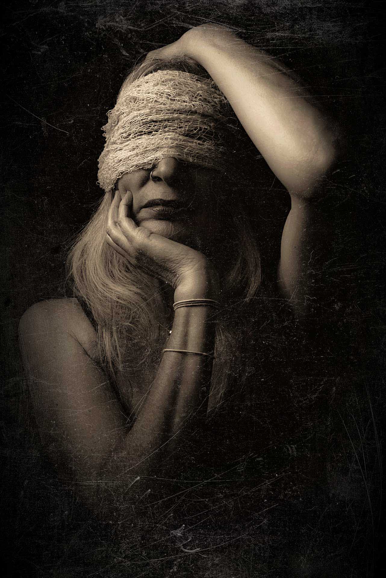 Moody portrait of a woman with her eyes covered by textured fabric, resting her head on her hand in low light.