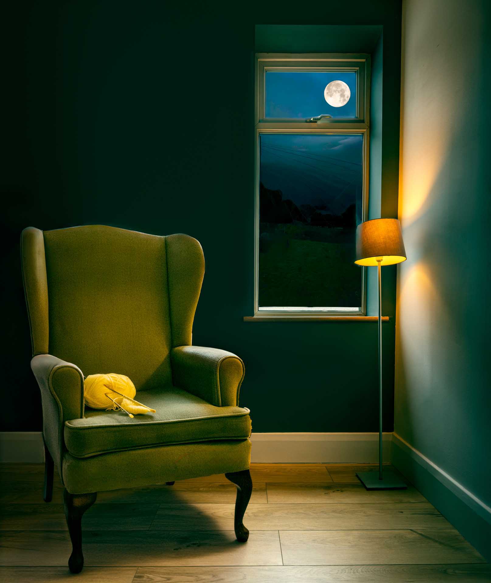 Cozy interior with an armchair, a ball of yellow yarn and knitting needles, a lit floor lamp, and a window showing a full moon at night.