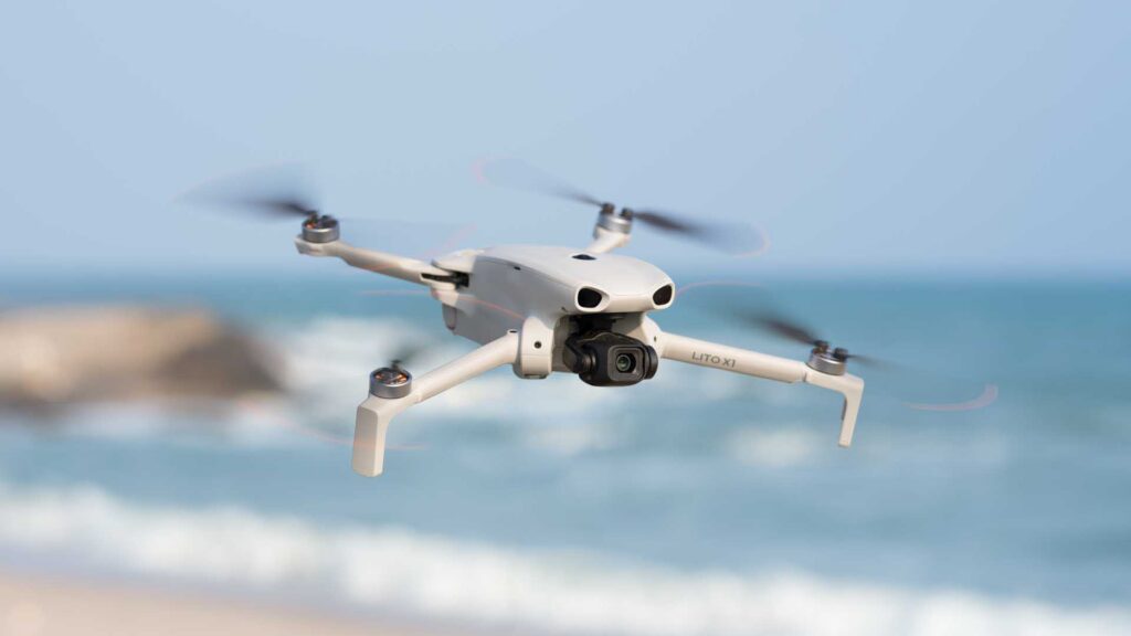 DJI Lito X1: a white quadcopter drone flying above a beach with ocean waves blurred in the background