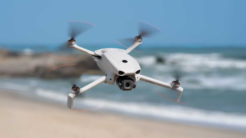DJI Lito 1: a white quadcopter drone flying above a beach with ocean waves blurred in the background