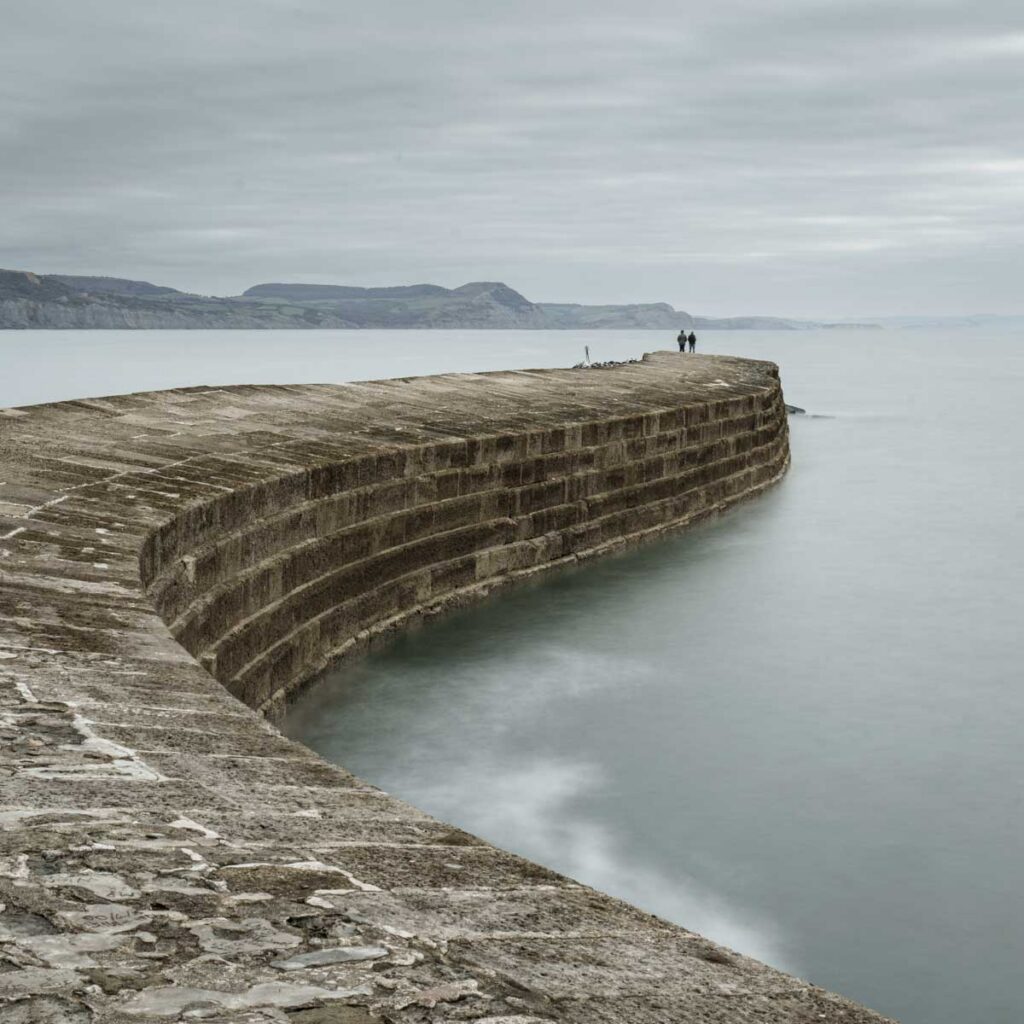Curved stone harbor wall extending into calm water under an overcast sky, with two small figures standing at the far end and distant cliffs across the bay.