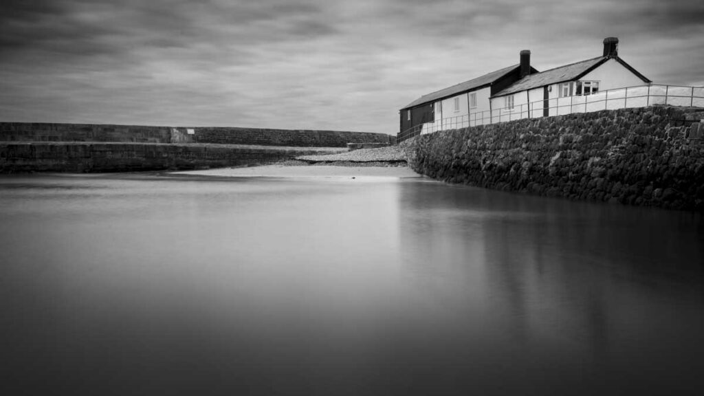 Black and white coastal scene with silky water, stone harbor walls, and a row of small buildings along the right side beneath a cloudy sky.