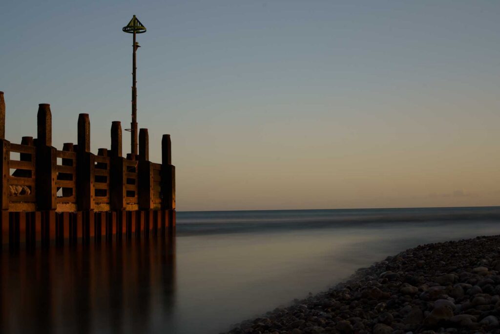 Wooden groyne structure and navigation marker at the edge of a beach, with smooth long-exposure sea and warm sunset tones on the horizon.
