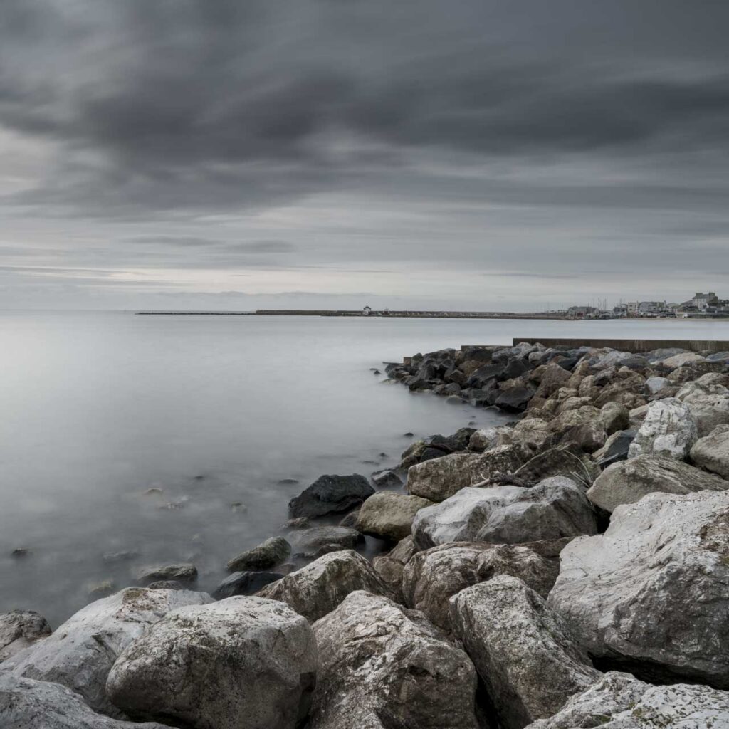 Rocky shoreline in the foreground leading to a calm, misty sea under heavy clouds, with a distant breakwater and small coastal town on the right.