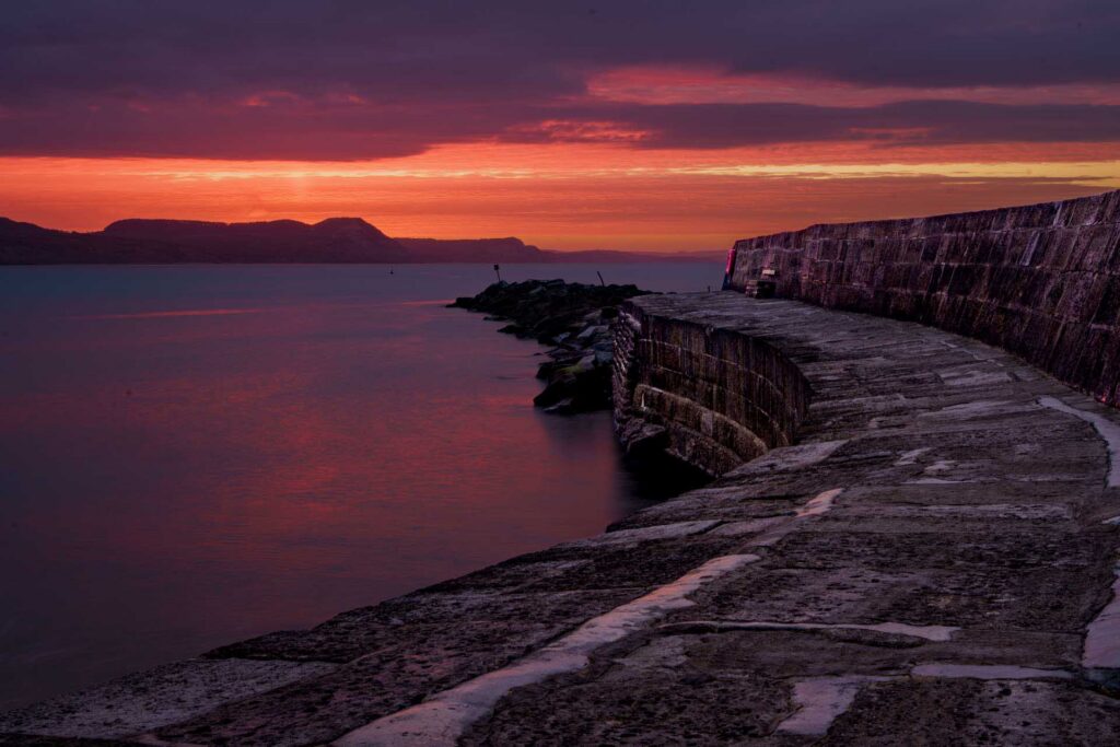 Curving stone pier at sunset with vivid red and orange sky reflecting on the water, and dark silhouettes of distant hills across the horizon.
