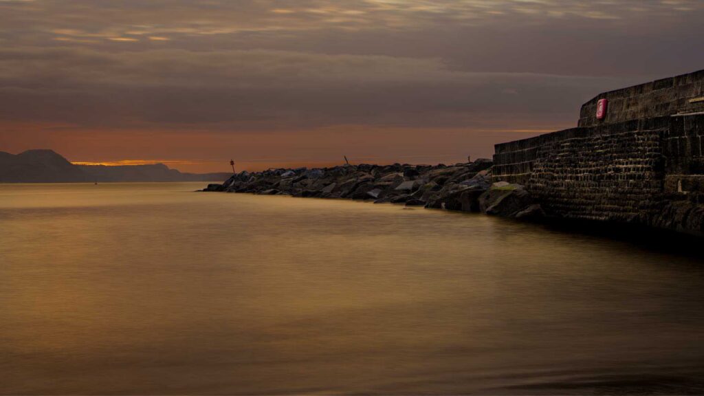 Harbor wall and rocky breakwater at golden hour, with smooth reflective water and a distant headland under a softly lit sky.