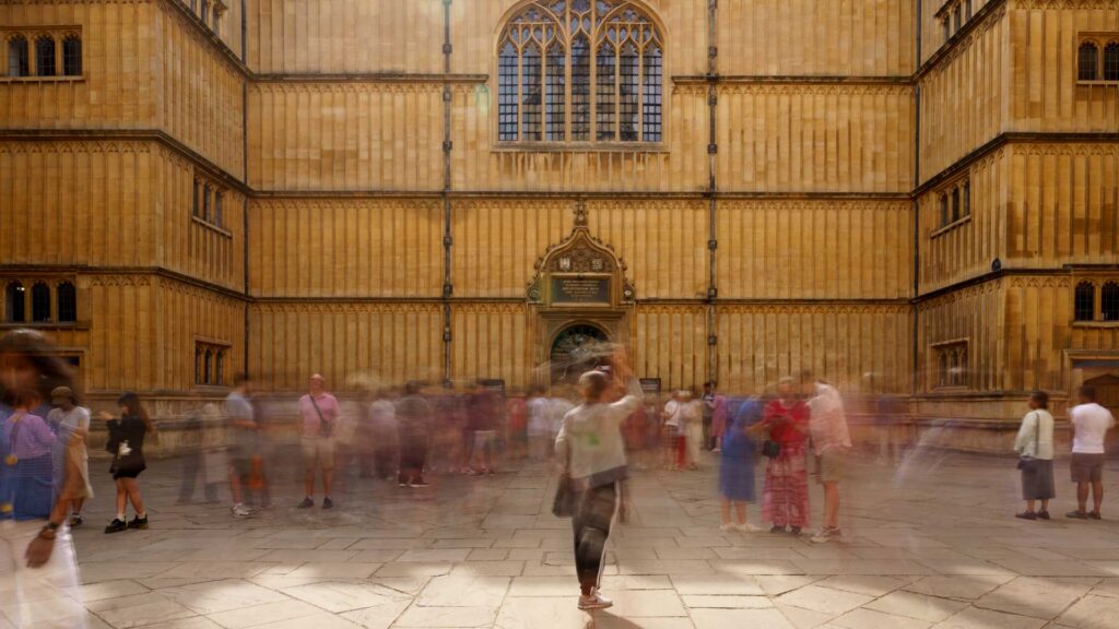 Blurred crowd of people moving through a stone courtyard in Oxford in front of a large historic building with tall arched windows, captured with long exposure to show motion.