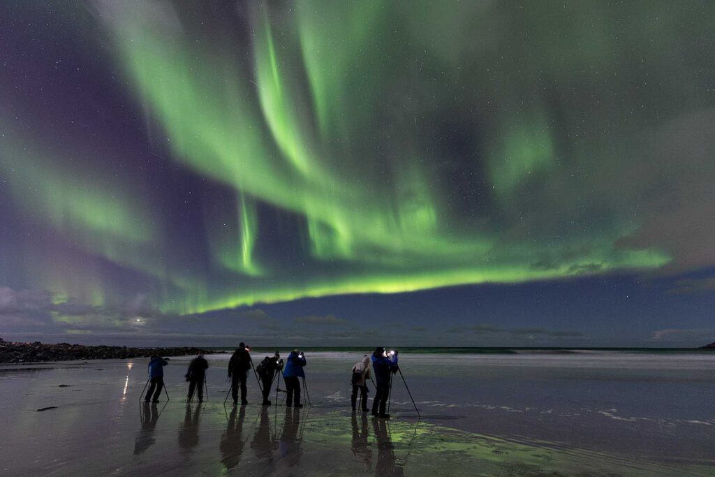 Group of photographers standing on a reflective beach at night capturing vivid green aurora borealis stretching across the sky.