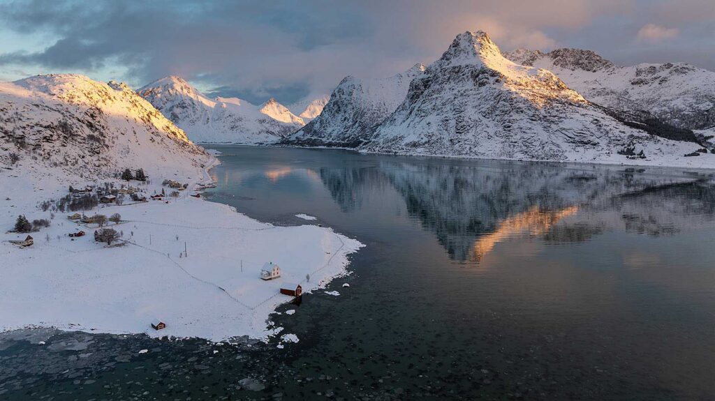 Wide fjord bordered by snowy peaks reflecting in still water, with scattered small houses along the shoreline at sunset light.