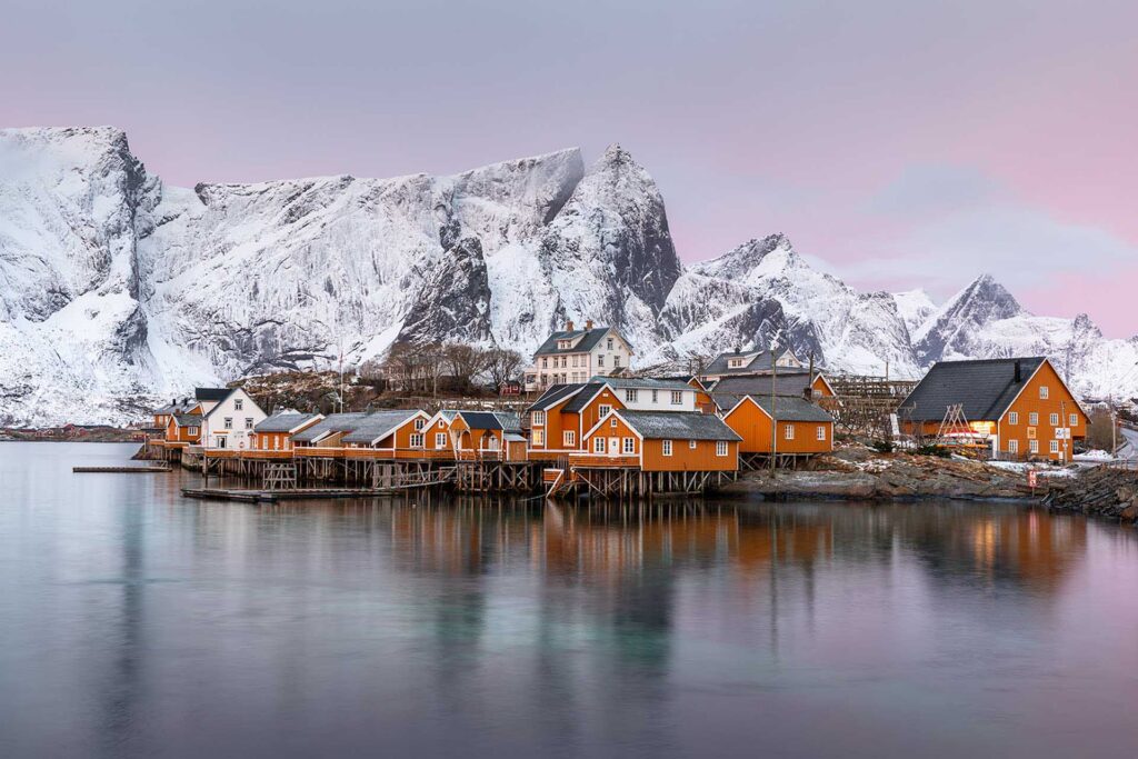 Row of orange fishing cabins on stilts along calm water with towering snow-covered mountains in the background under a soft pink sky.
