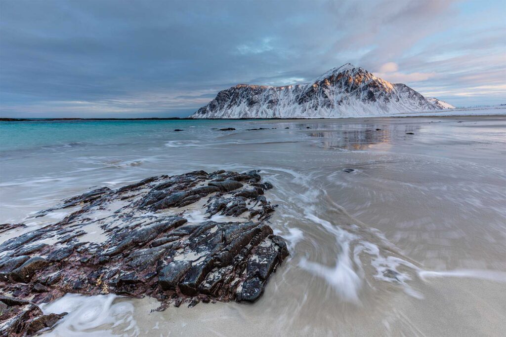 Rocky beach with swirling water patterns leading toward a distant snow-covered mountain under a cloudy blue sky.