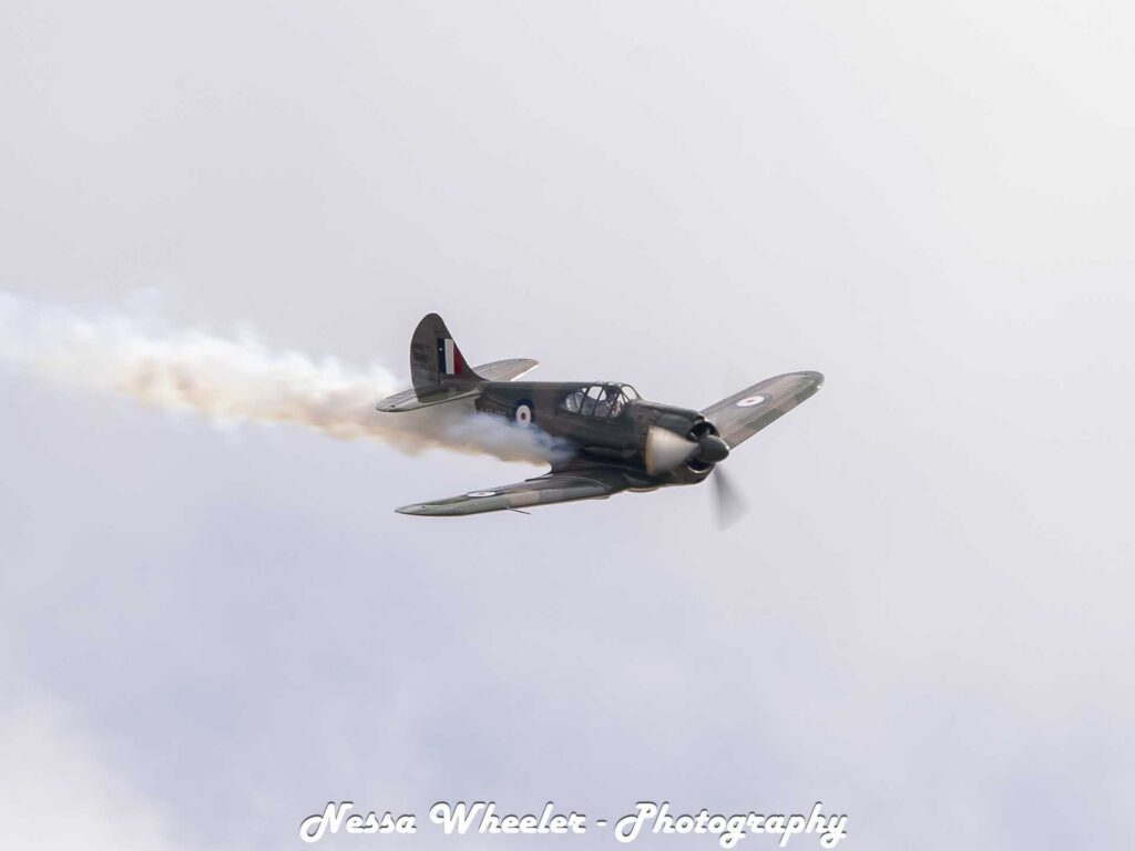 Historic propeller aircraft flying through a pale sky with a smoke trail, with “Nessa Wheeler - Photography” overlaid at the bottom.
