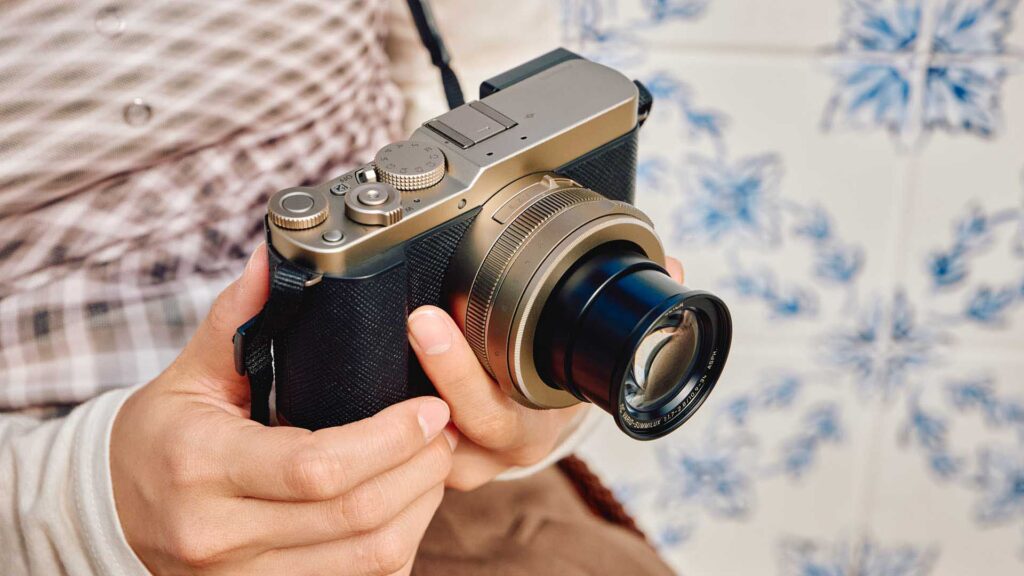 Compact Panasonic Lumix camera with lens extended, held in both hands against a softly patterned background.