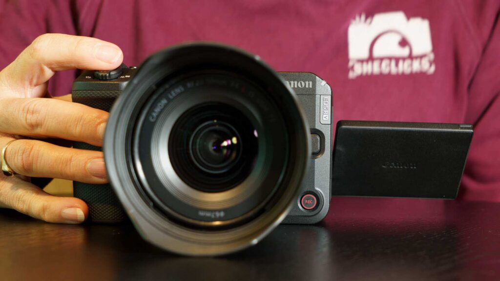 Front-facing view of the Canon EOS R6 V with the RF 20-50MM F4L IS USM PZ lens attached and the side flip screen opened outward. A hand steadies the camera from the left side.