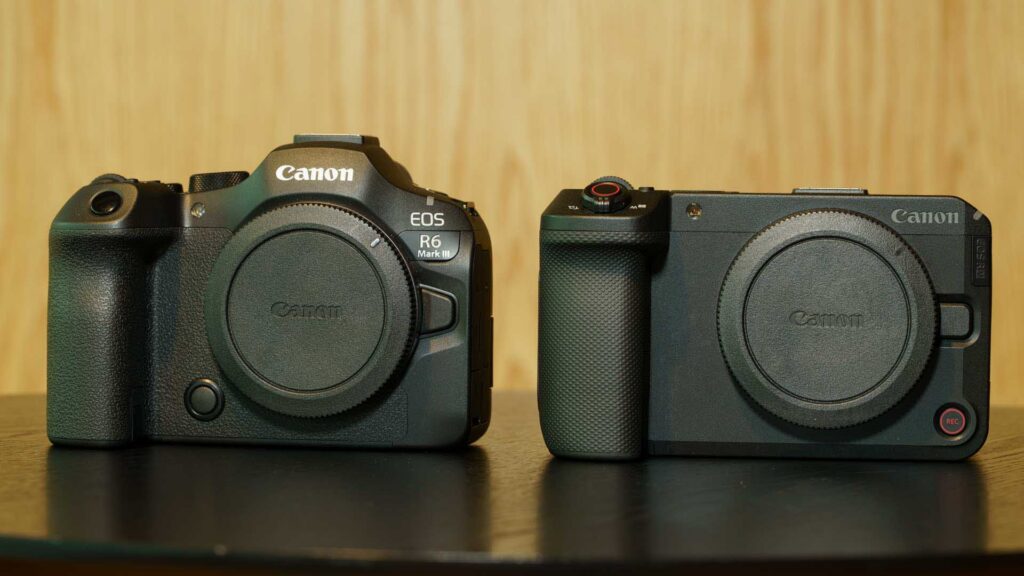 Front view comparison of the Canon EOS R6 Mark III and Canon EOS R6 V camera bodies side by side on a table. The EOS R6 Mark II has a larger DSLR-style grip and electronic viewfinder hump, while the EOS R6 V has a more compact cinema-style body.