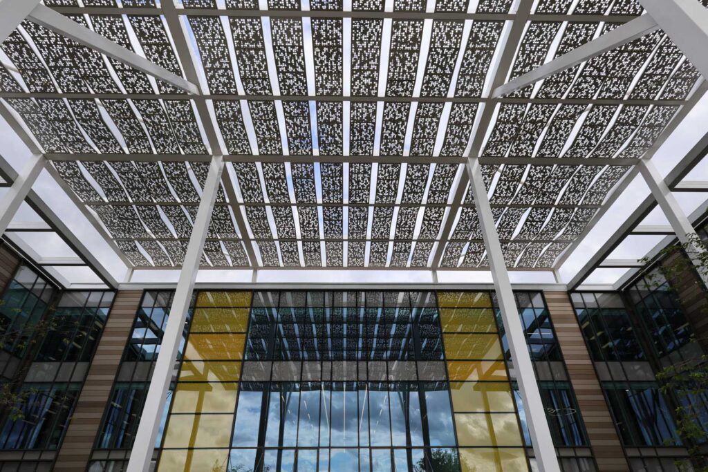 Modern building entrance with large glass windows beneath a patterned metal canopy supported by white columns, viewed from below.