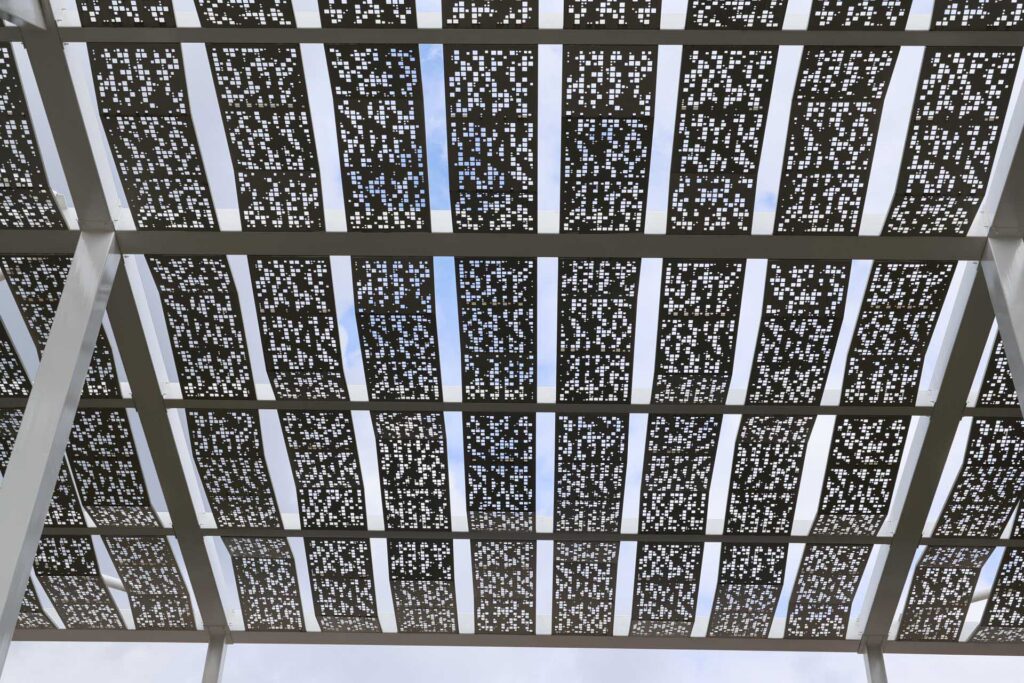 Upward view of a geometric metal canopy with decorative perforated panels and white support beams against a cloudy sky.