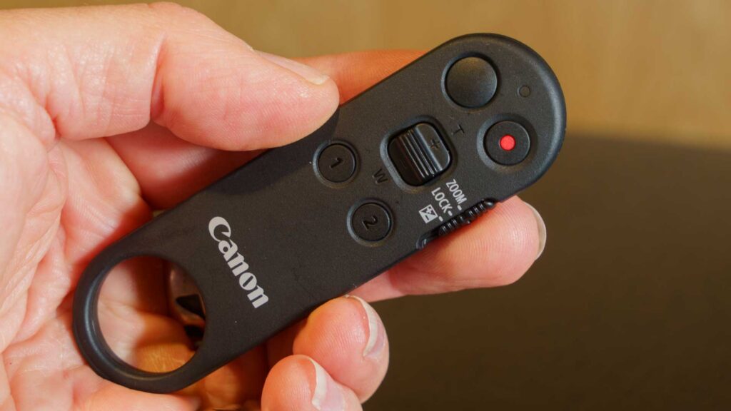 Close-up of the Canon BR-E2 remote control showing zoom, record, and custom buttons held between two fingers.