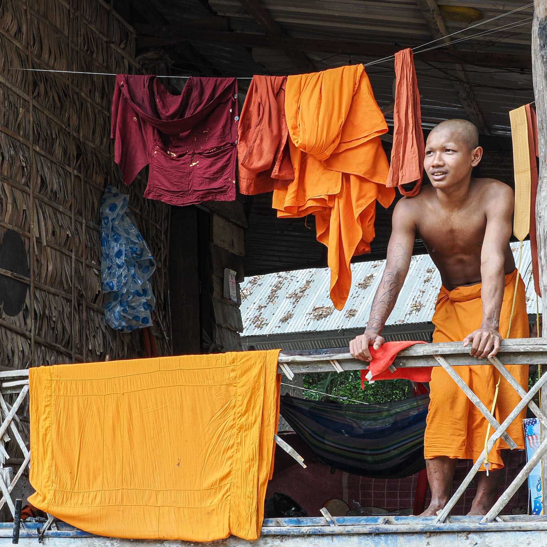 Young Buddhist monk standing on a balcony beside orange robes hanging to dry.