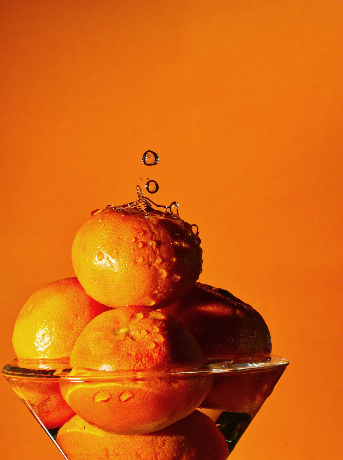 Stacked oranges in a glass bowl with water droplets splashing against an orange background.