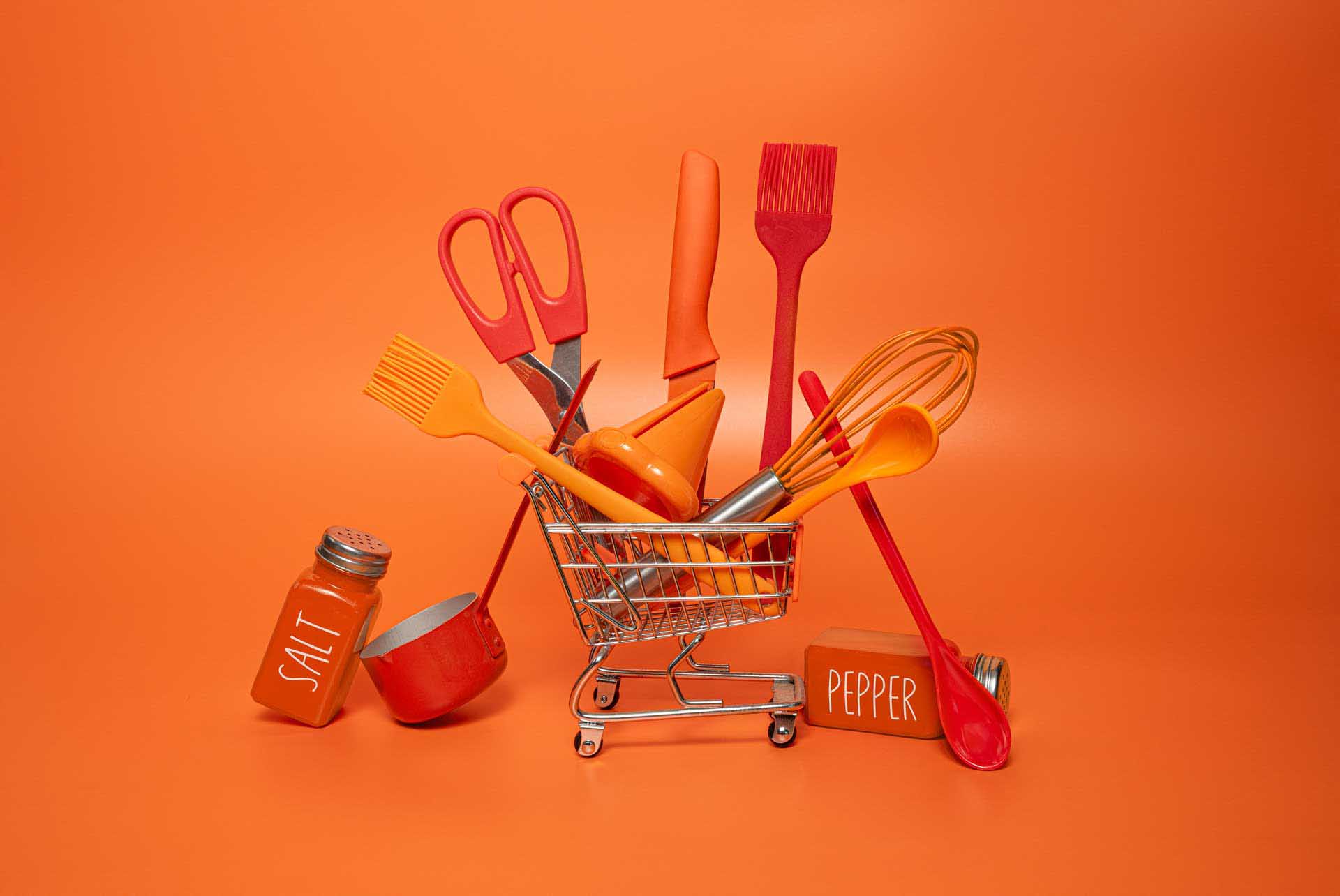 Mini shopping trolley filled with orange kitchen utensils against an orange backdrop.