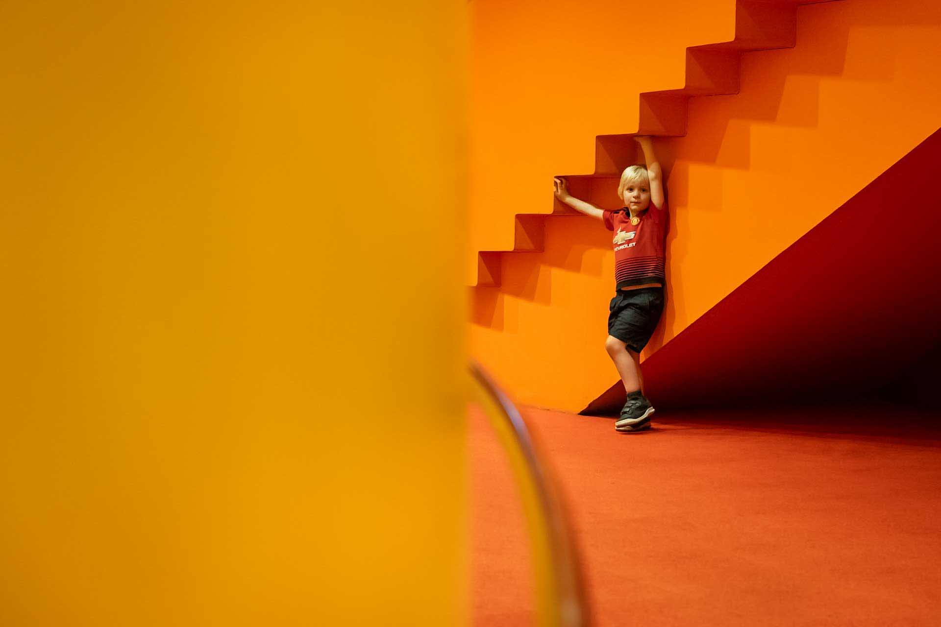 Child standing beneath an orange staircase in a minimalist interior.
