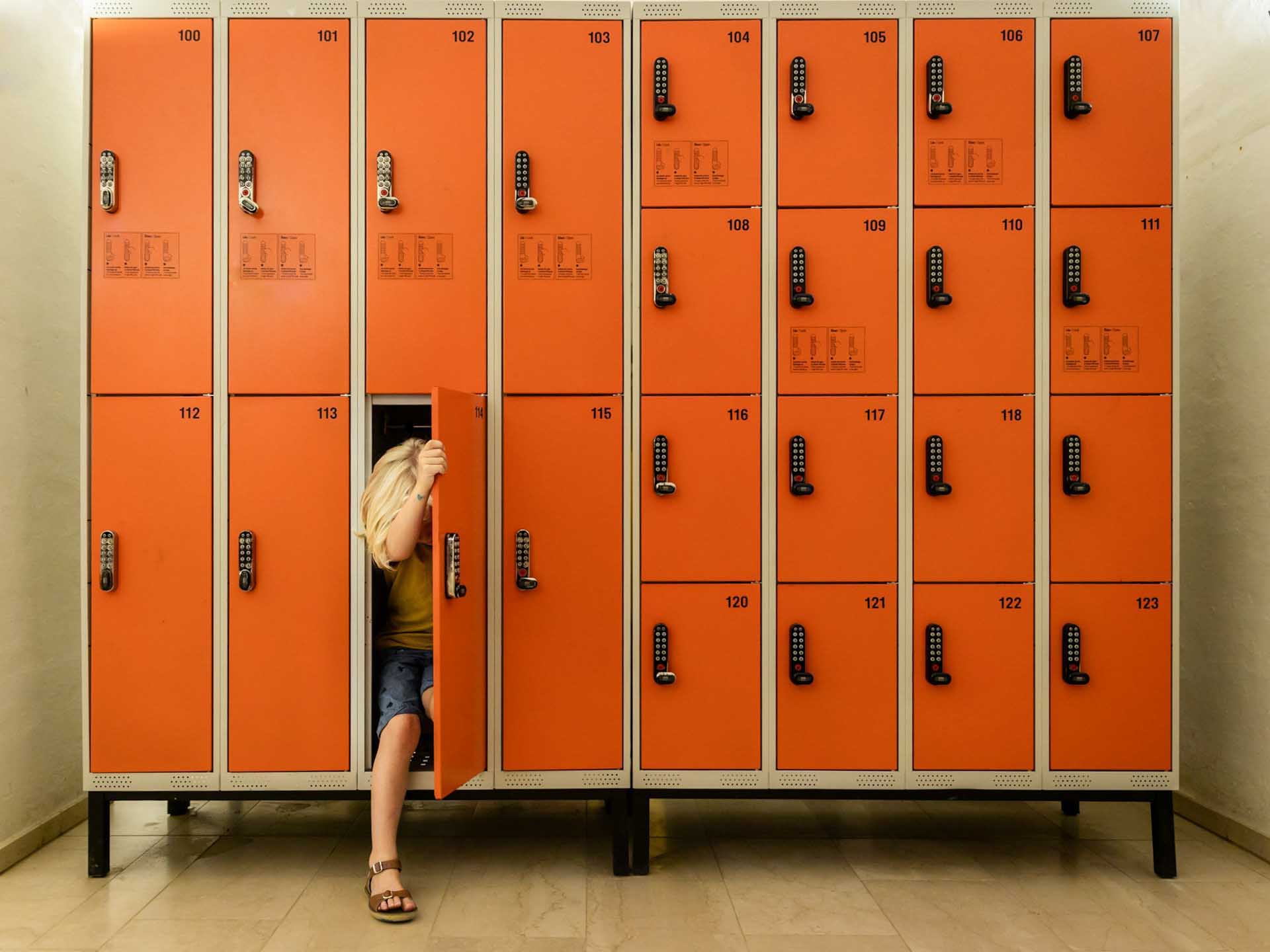 Child stepping out from an orange locker in a wall of numbered lockers.