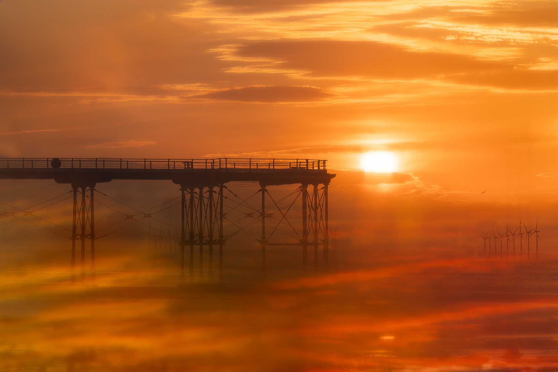 Silhouetted pier extending over water at sunset with orange reflections and distant wind turbines.