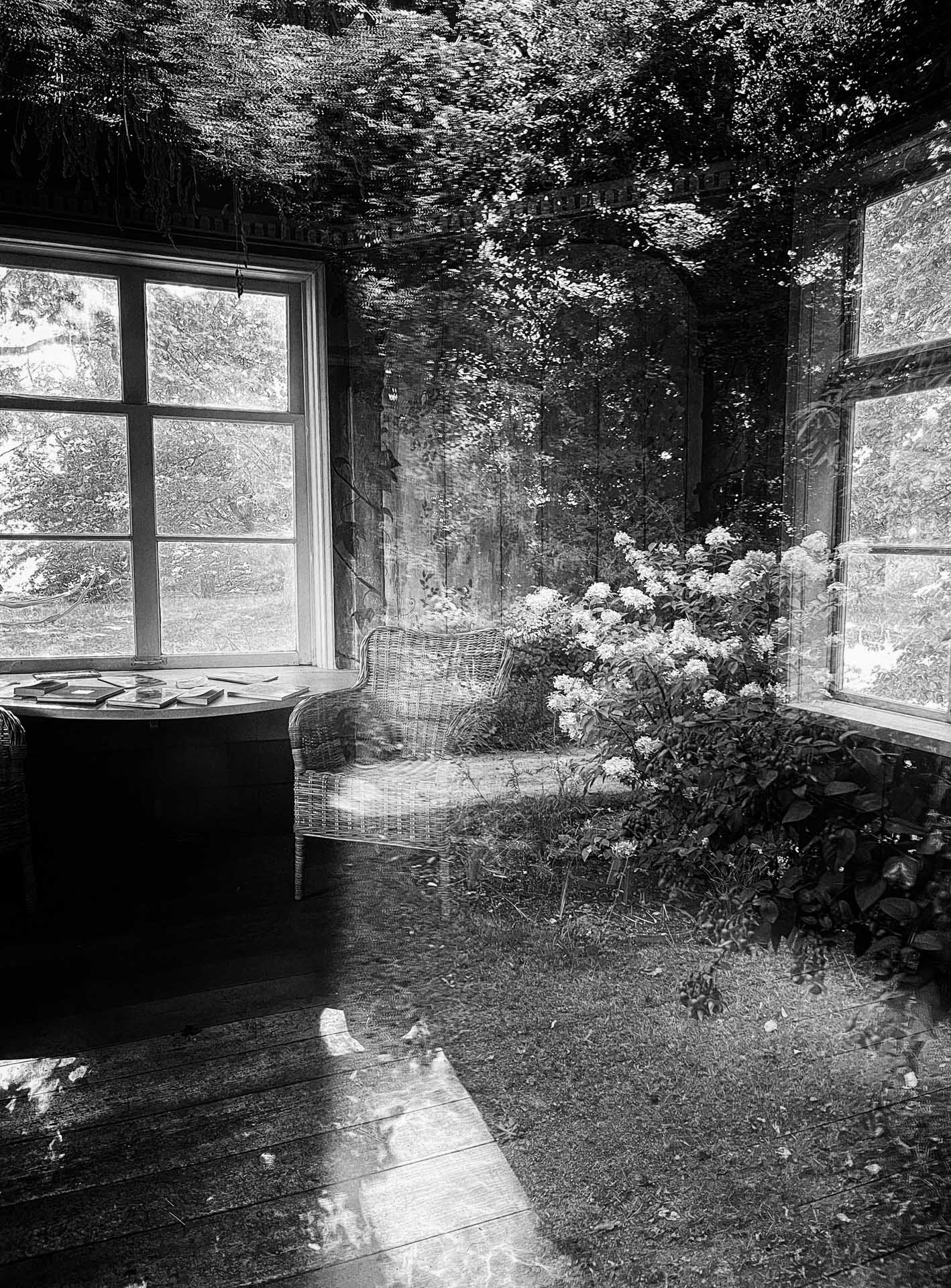 Black-and-white double-exposure image combining a sunlit room with wicker chairs and windows with an outdoor garden of trees and flowering shrubs.