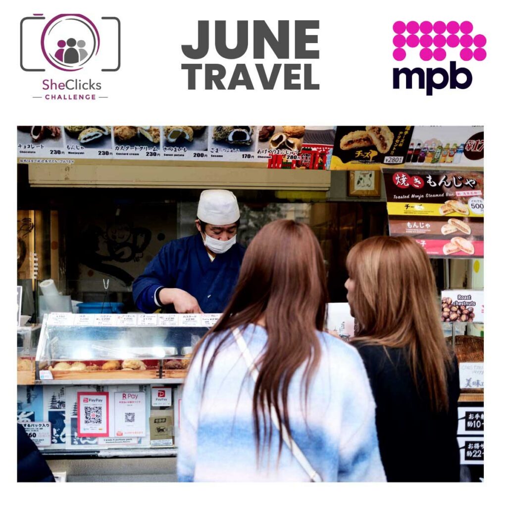 Promotional graphic with SheClicks Challenge, June Travel, and MPB logos across the top. Below, a street food vendor wearing a mask serves customers at a Japanese snack stall. Menu boards display pastries and prices in yen, while two women stand at the counter ordering.
