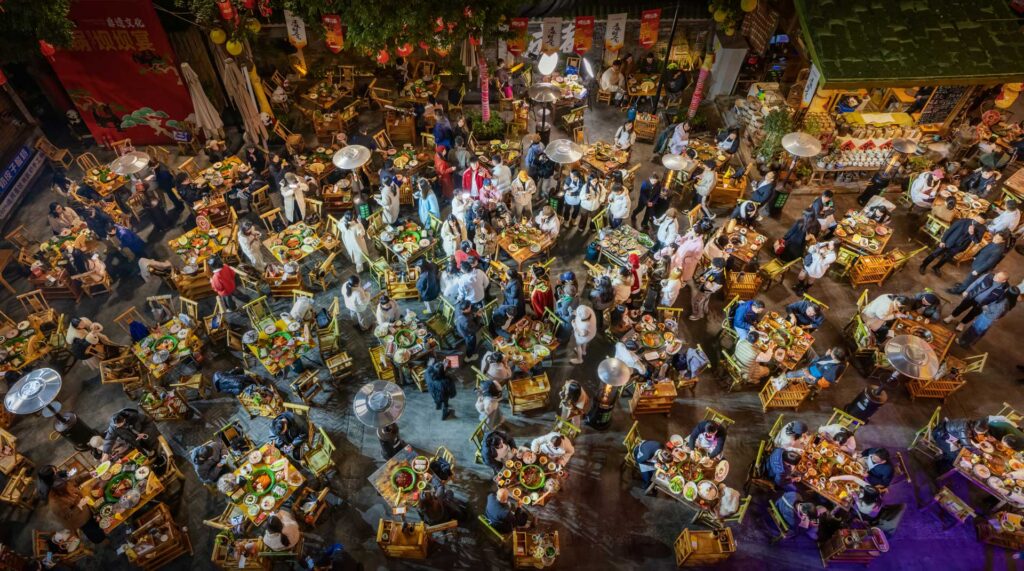 Overhead view of a crowded outdoor restaurant at night, with diners gathered around dozens of brightly lit tables.