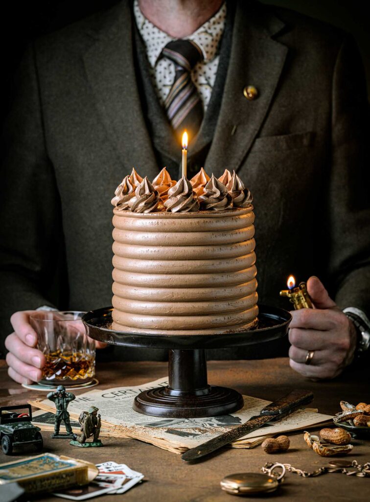 Person in a suit holding a tall chocolate cake with a single lit candle, surrounded by keepsakes, cards, and a glass of whiskey.
