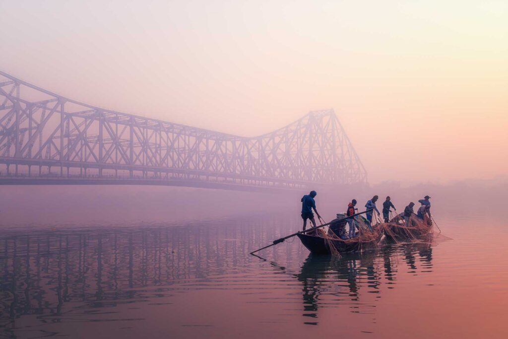 Fishermen standing in narrow boats on calm water at dawn, with a large steel bridge fading into the mist behind them.