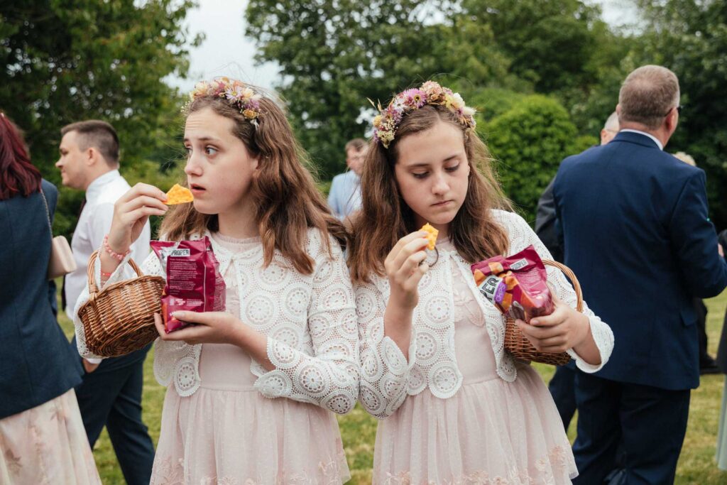 Two girls wearing flower crowns and matching dresses eat chips while holding wicker baskets at an outdoor gathering.