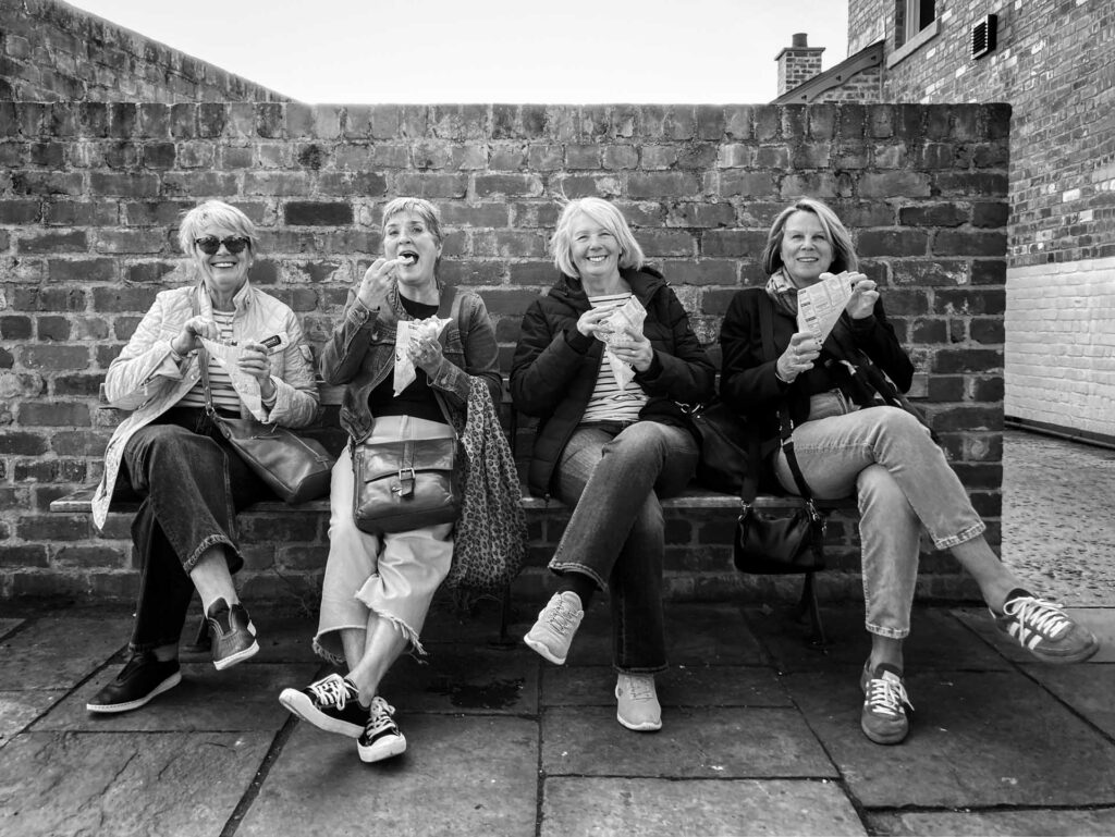 Four women sitting on a bench against a brick wall, smiling and eating snacks in a black-and-white photograph.