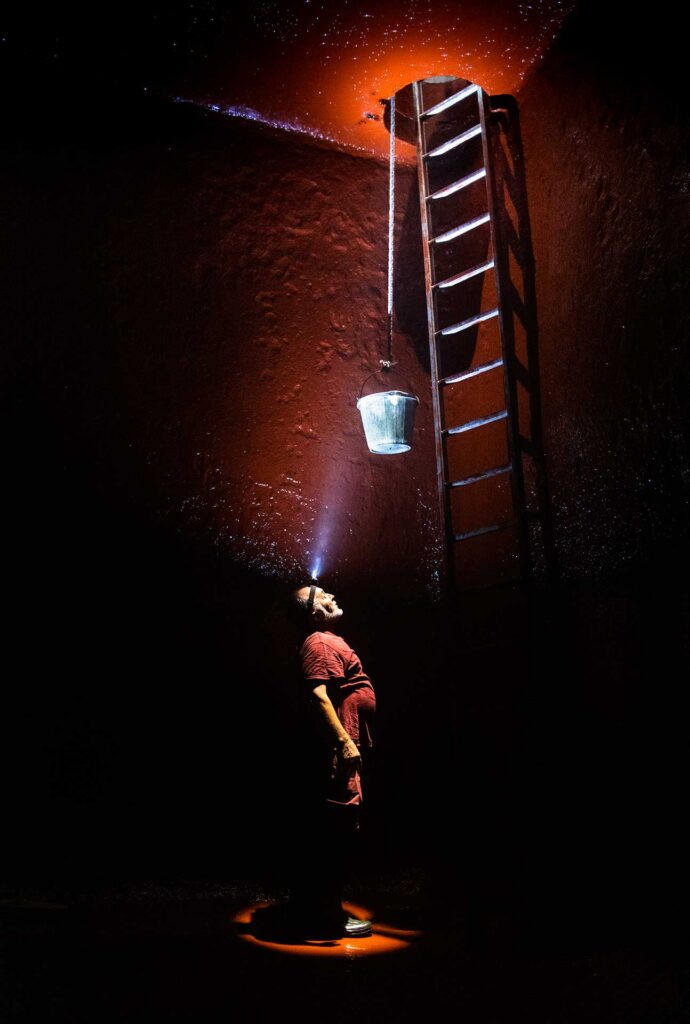Worker shining a headlamp upward inside a dark tank, illuminating a ladder and a hanging bucket.