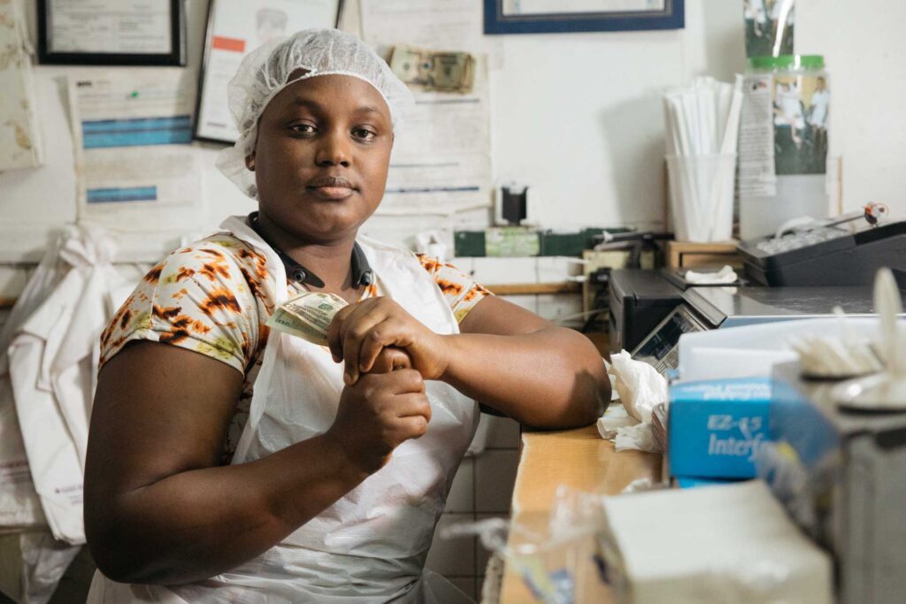 Food-service worker seated at a counter, holding a folded dollar bill in a small office or kitchen workspace.