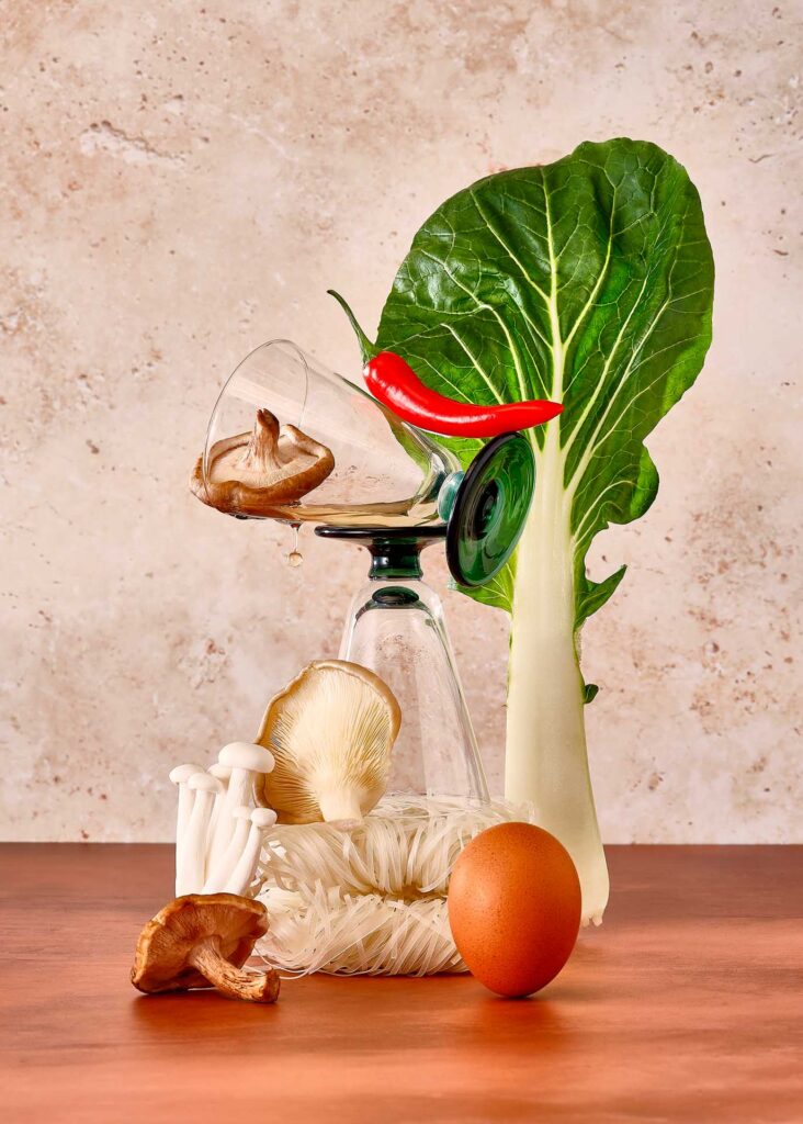 Still life of bok choy, mushrooms, chili pepper, noodles, and an egg balanced around stacked glassware.