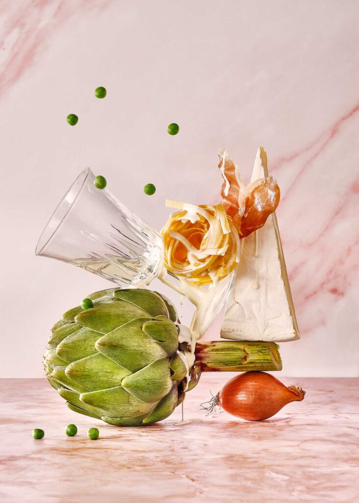 Artichoke, wedge of soft cheese, pasta nest, peas, and onion arranged in a playful balancing still life.