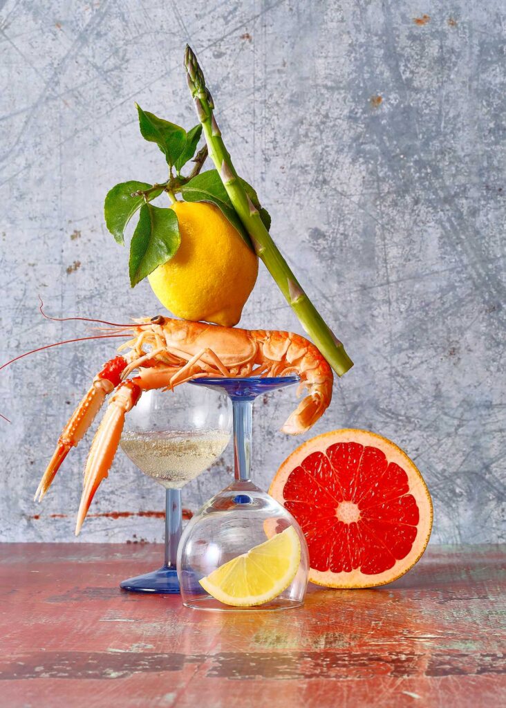 Langoustine, lemon, asparagus, grapefruit, and wine glasses stacked in a colorful seafood still life.